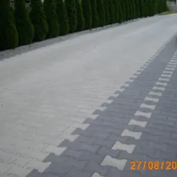 Szeroki podjazd wyłożony jasnoszarą kostką brukową o nieregularnym kształcie, z dekoracyjnym pasem ciemnoszarej kostki z jasnymi wstawkami po prawej stronie, obrzeża z zielonych krzewów i wysokich...